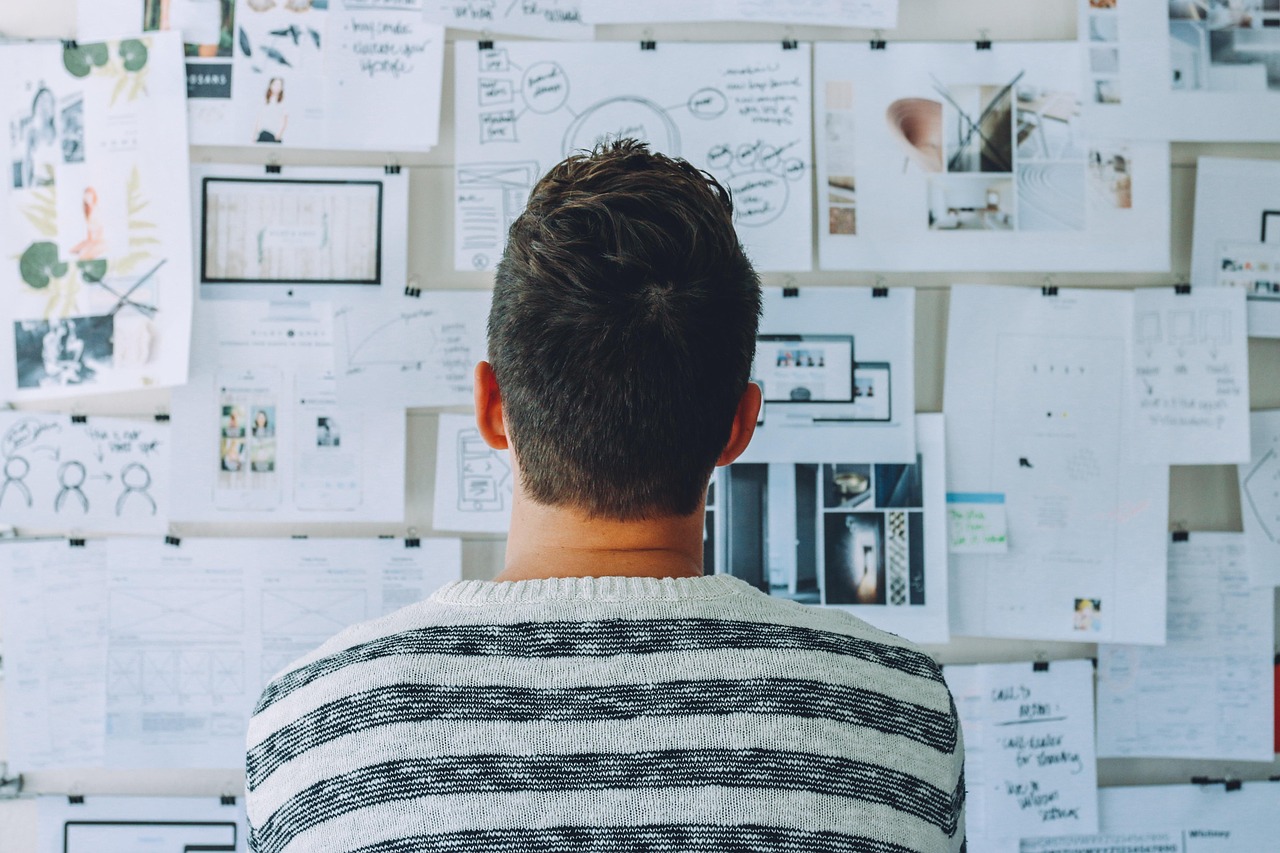 A man seen from behind studies a wall of pinned sketches, diagrams and notes.